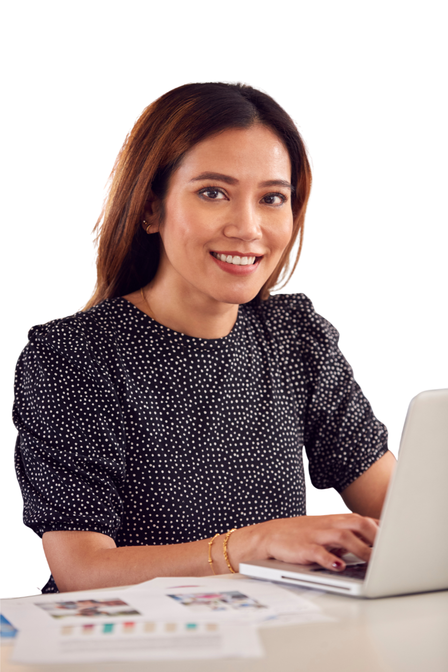 A smiling woman with medium-length brown hair working on a laptop at a white desk with papers and photographs, wearing a black polka dot blouse.