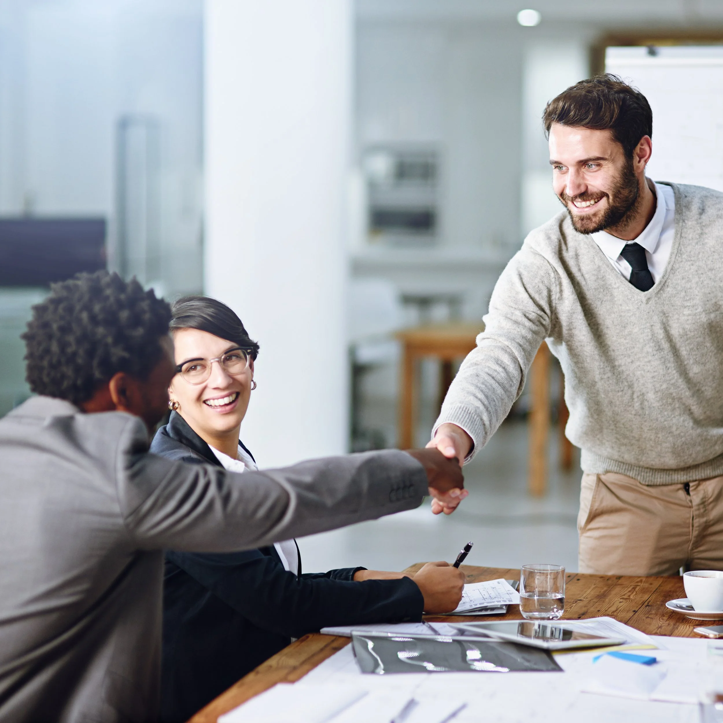 A man with dark hair, a beard, and a gray sweater is smiling and shaking hands with a woman. They are sitting at a wooden table in a bright office space, possibly engaging in a business meeting or interview, with documents and a glass of water on the table.