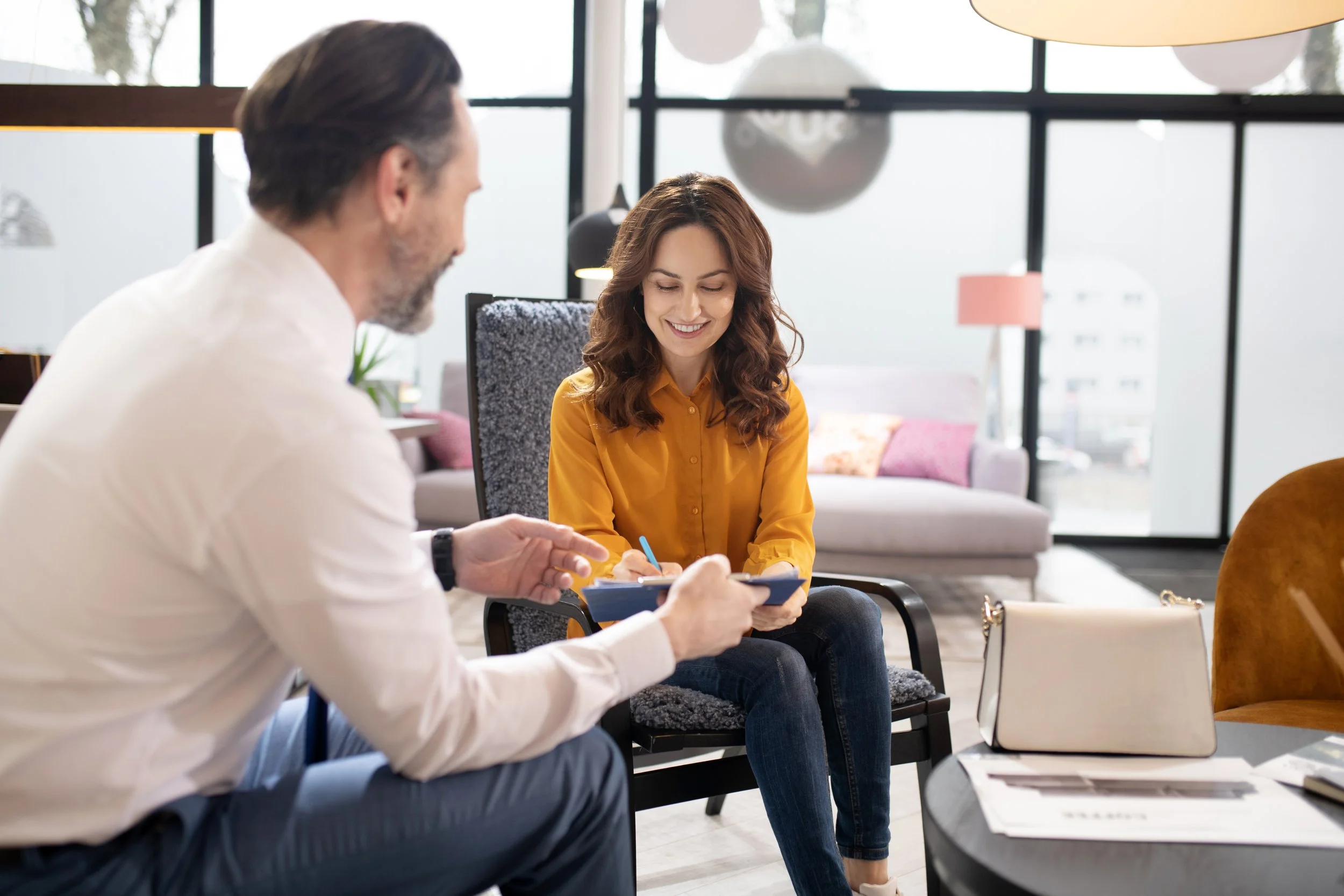 Two men and a woman sit in a bright, modern lounge area engaged in a discussion, with the woman smiling and taking notes.