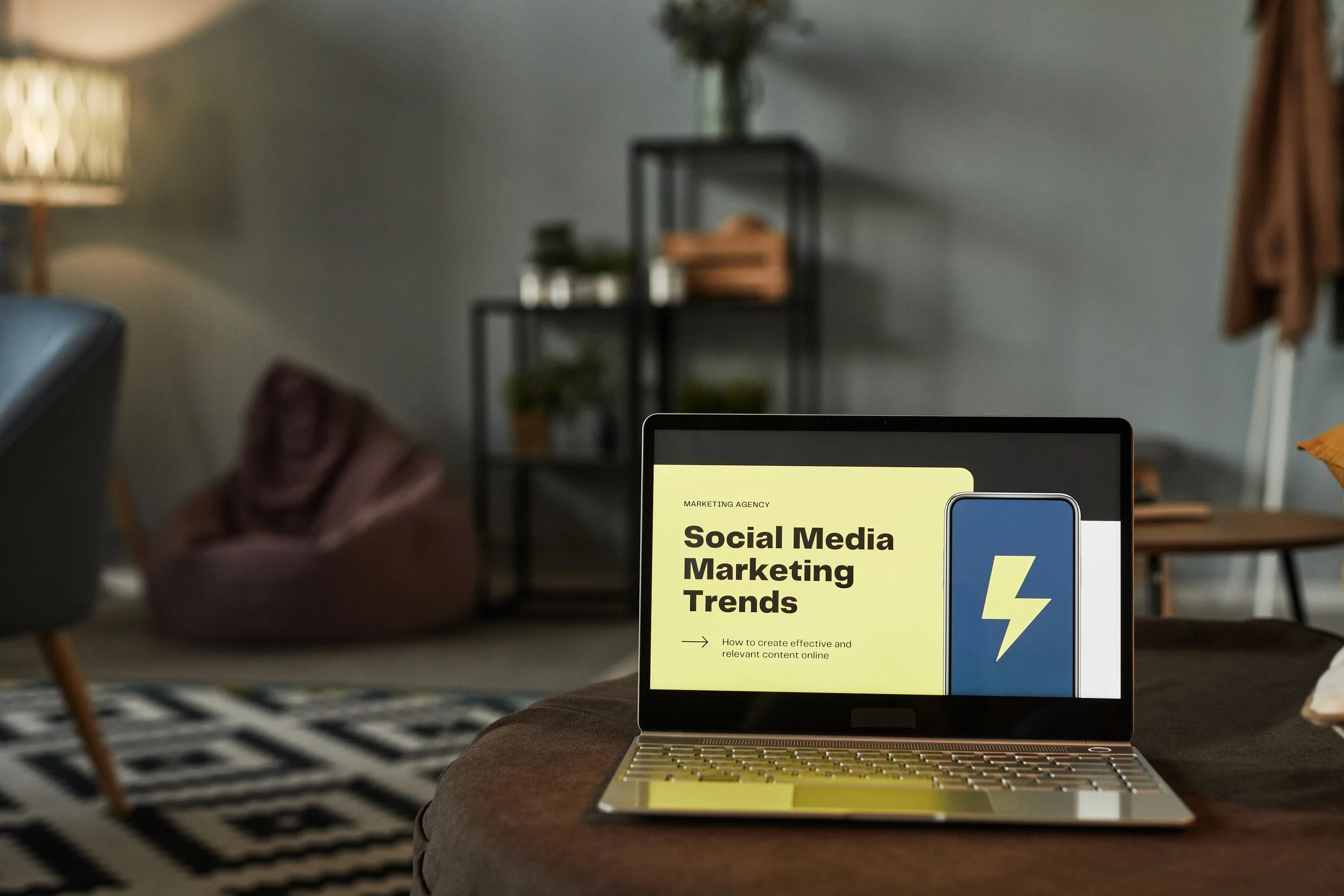Close-up of a laptop displaying a presentation slide titled 'Social Media Marketing Trends' in a cozy, modern living room with a black bookshelf, green plants, a lamp, and a brown chair in the background.