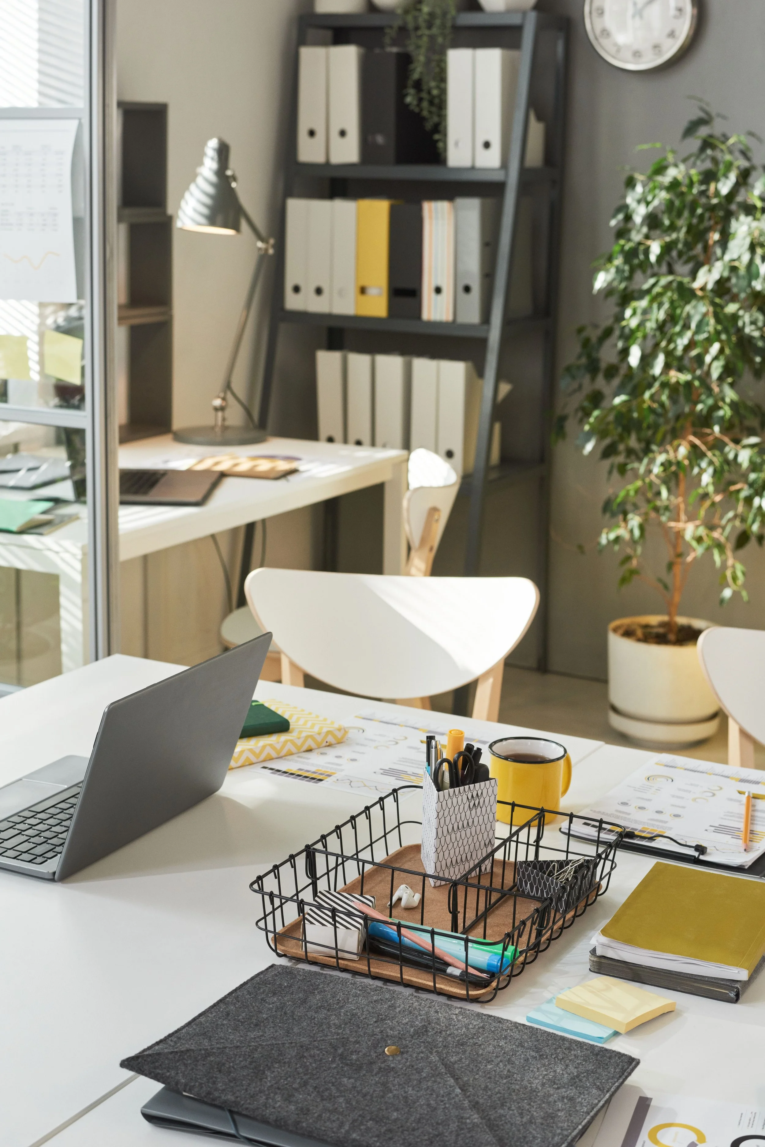 An organized office workspace with a white desk, laptop, various stationery items, and a wire desk organizer in the foreground. In the background, there is a bookshelf with binders and a potted plant, a white chair, and a large clock on the wall.