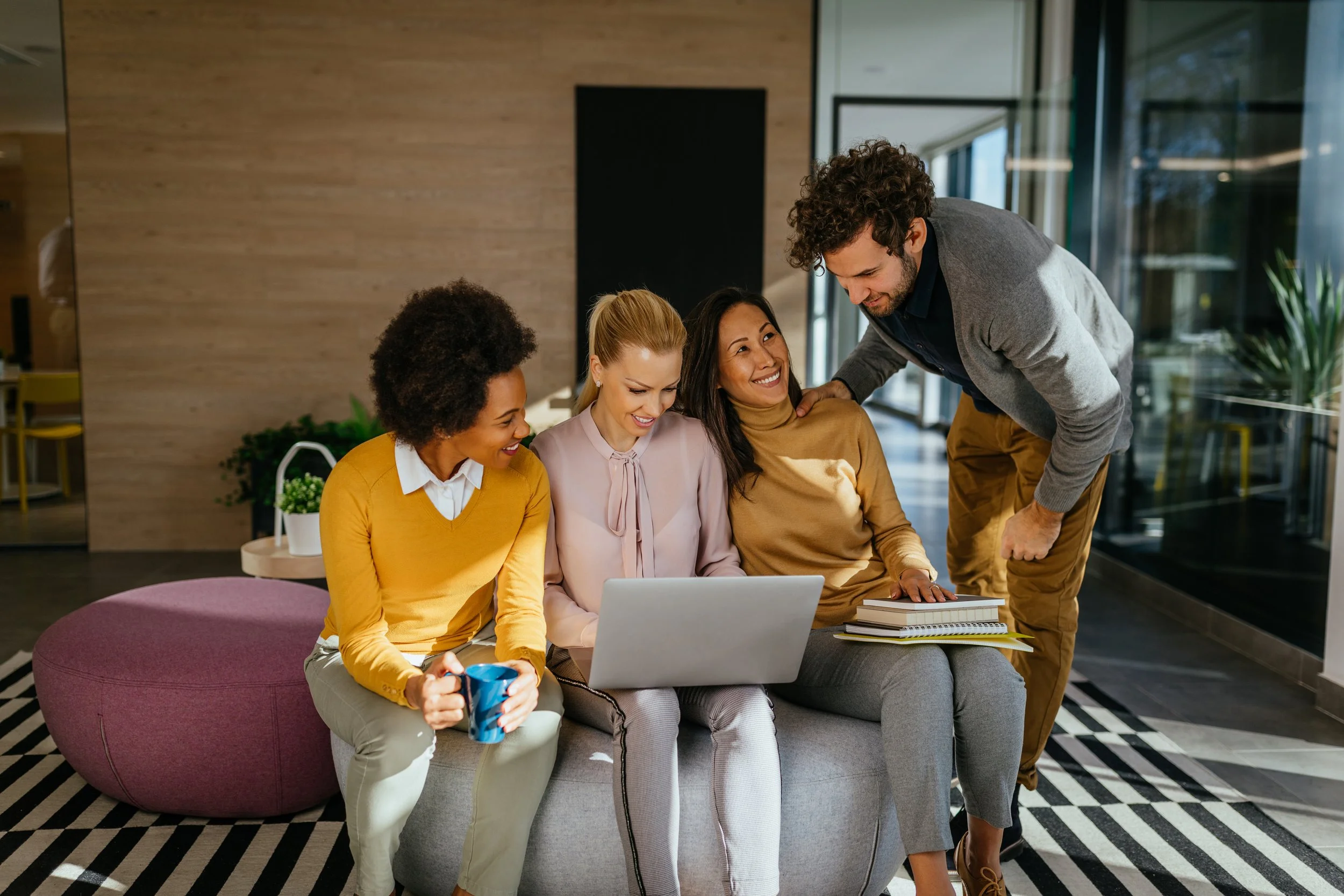 Four women and one man gathered around a laptop in a modern office space, smiling and looking at the screen, with notebooks and a coffee mug on the table.