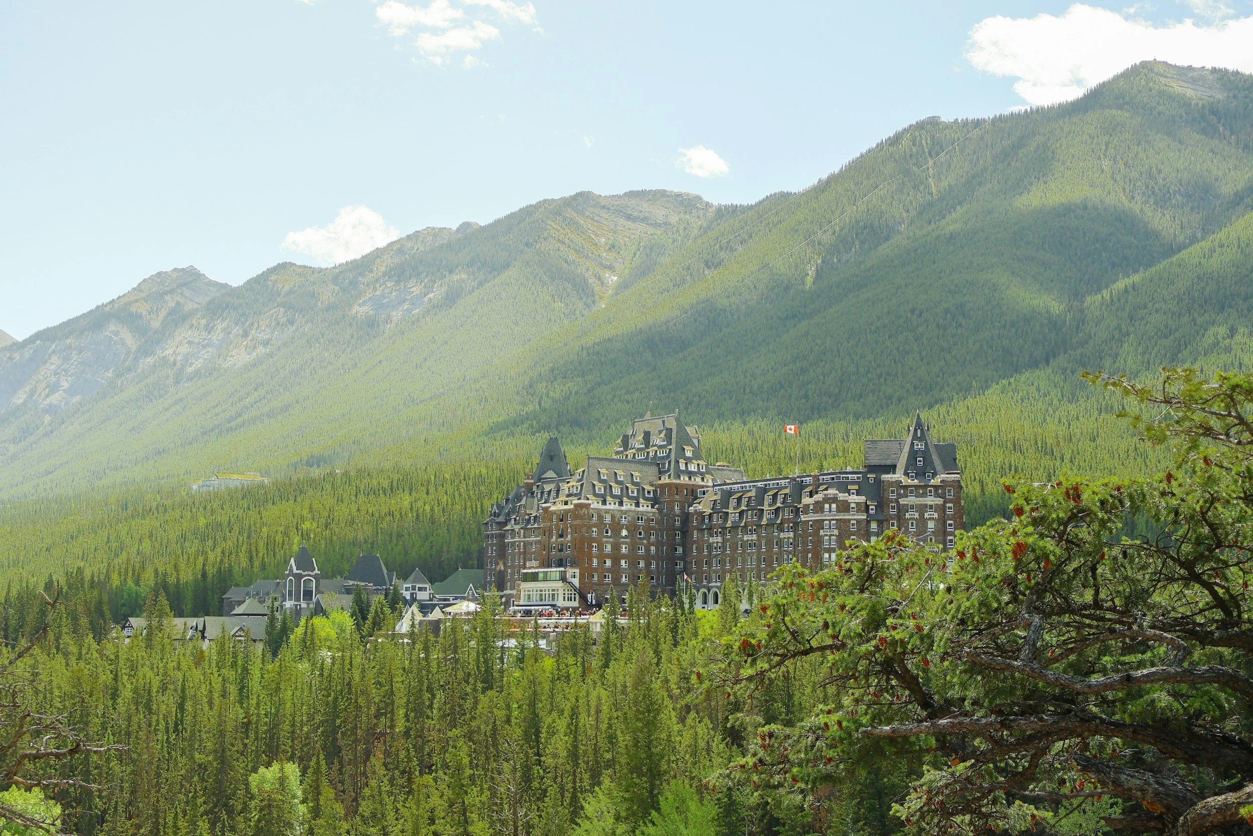 A large hotel building with dark turrets surrounded by green trees, situated at the base of green mountains with a cloudy sky above.