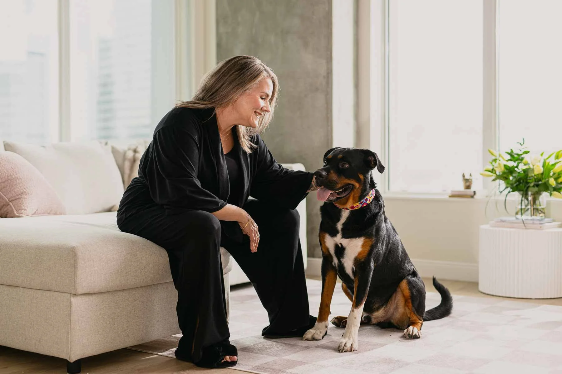 An older woman with grey hair sitting on a beige sofa, smiling and petting a happy black and brown dog with a colorful collar, in a bright living room with large windows.