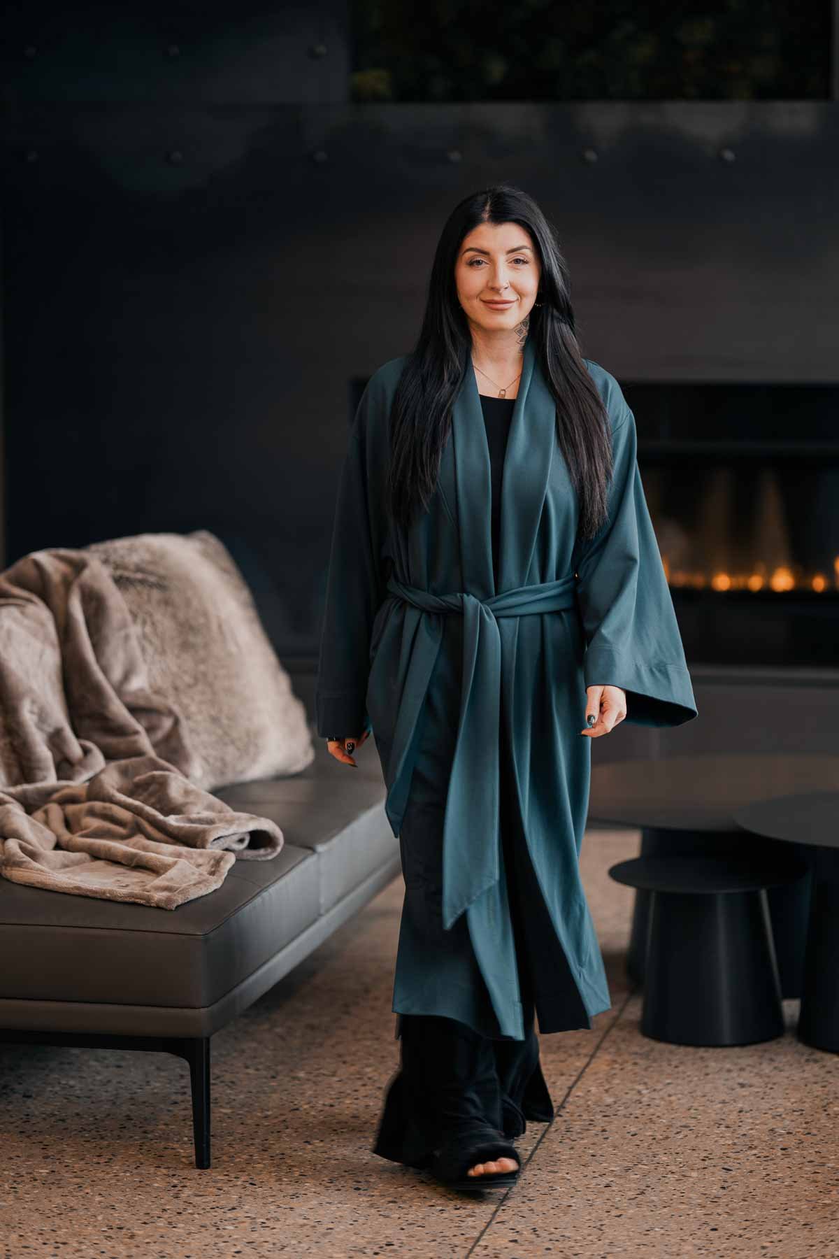 A woman with long black hair, wearing a teal silk robe over black clothing, walking indoors in front of a fireplace with a beige couch and furry pillows.