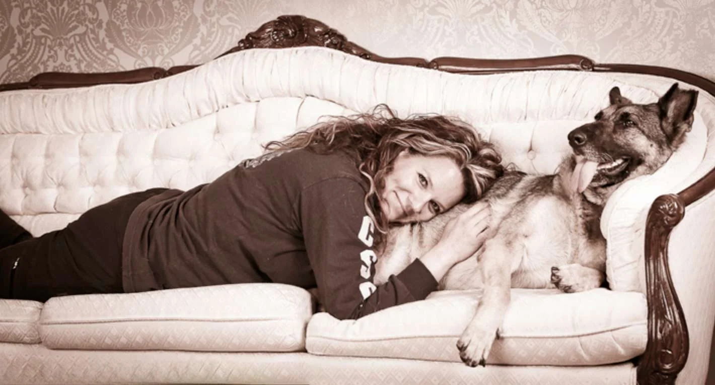 A woman lying on her side on a vintage sofa with a light-colored upholstery, resting her head on a large dog that is lying on the sofa, with its tongue out and smiling.