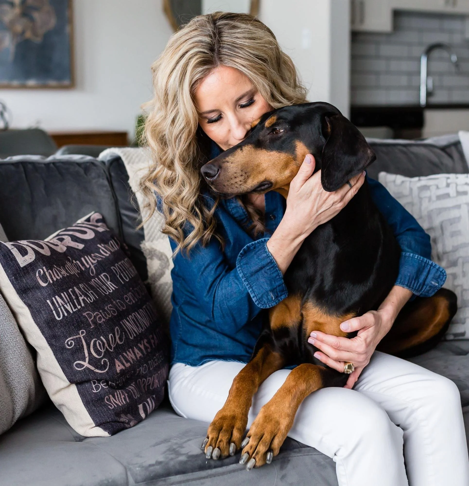 A woman with curly blonde hair hugging a large black and tan dog on a sofa in a cozy living room.