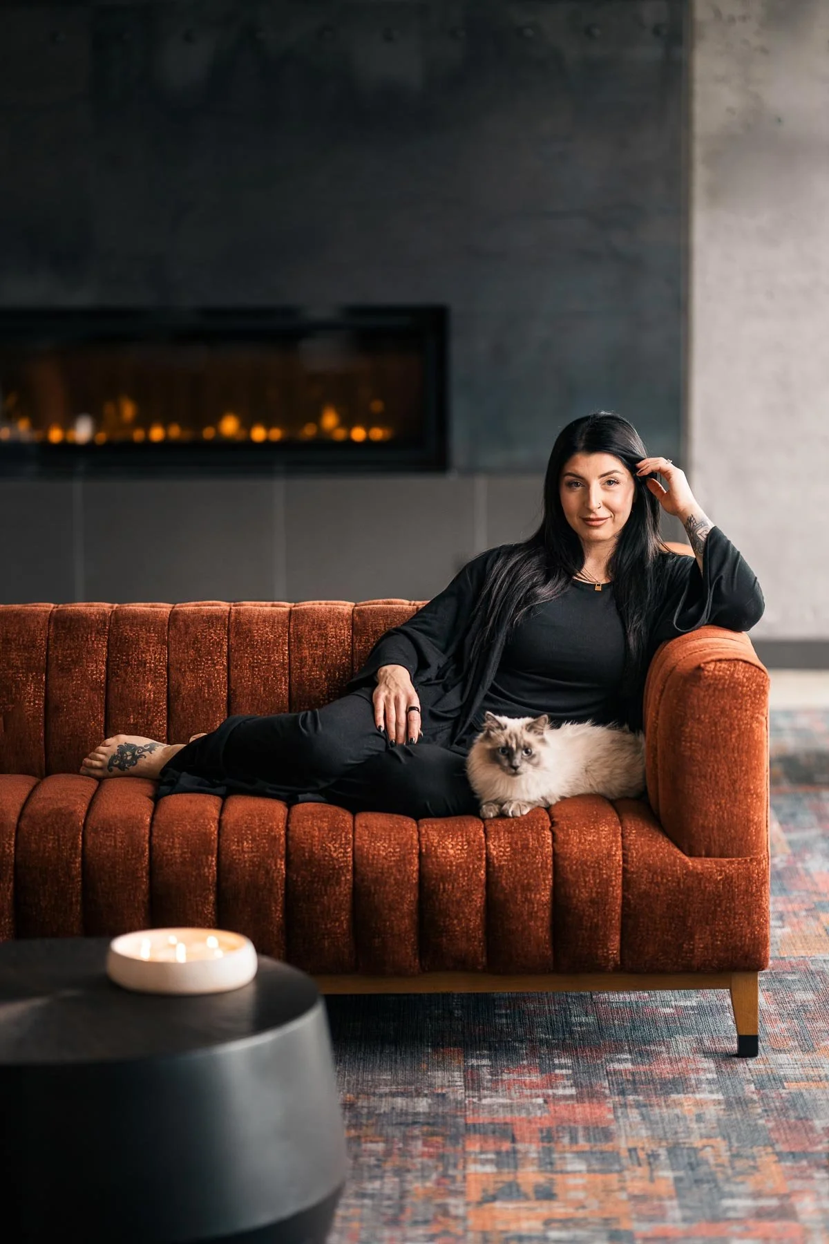 A woman with long black hair, wearing black clothing, reclines on an orange velvet sofa with a fluffy Siamese cat beside her. She is in a modern, cozy living room with a fireplace in the background, a round black coffee table in the foreground, and a patterned rug on the floor.