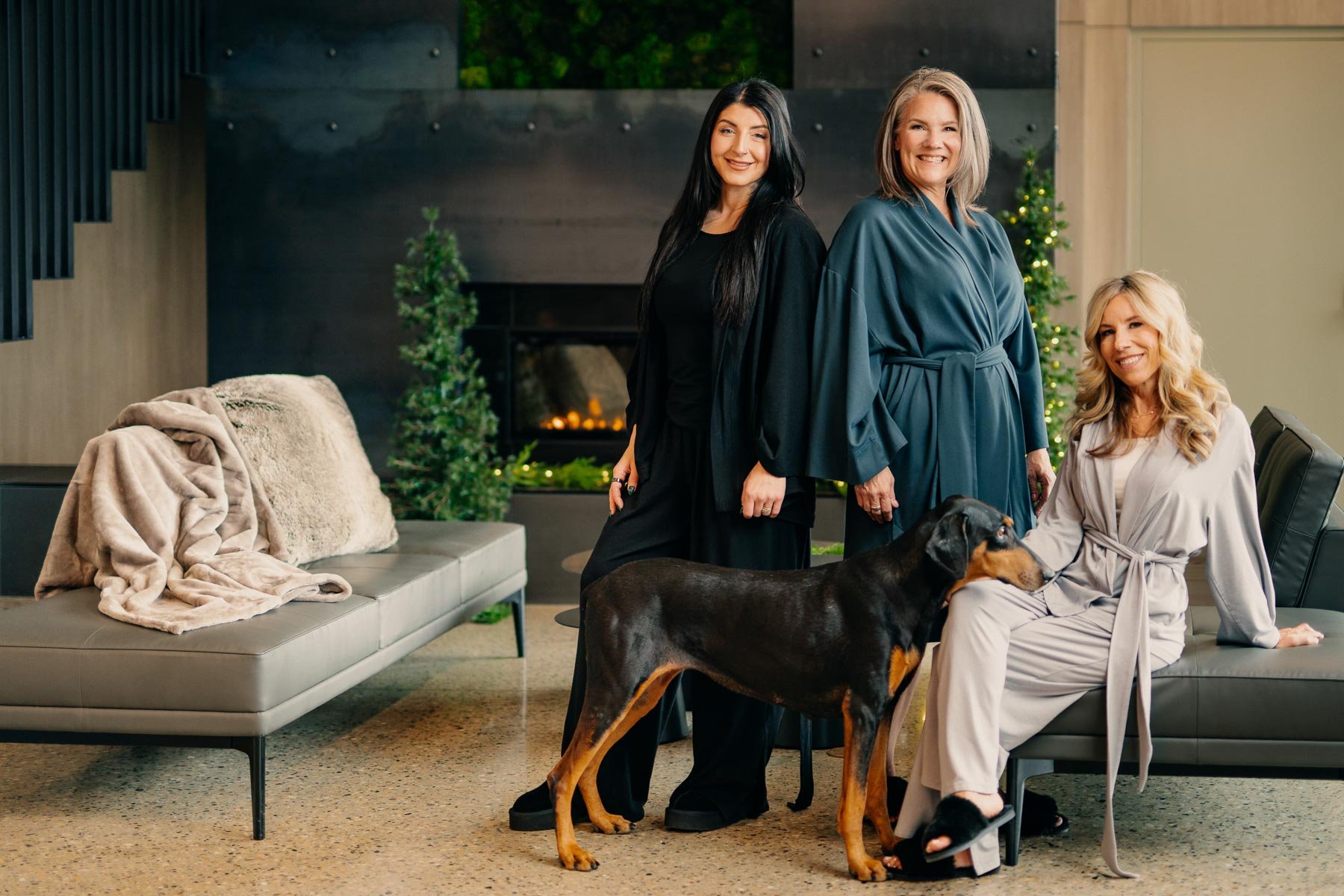 Three women and a dog in a modern living room decorated for Christmas. One woman is sitting on a couch, another is standing, and the third is standing slightly behind. The dog is a large black and tan breed. There are Christmas trees and a fireplace in the background.