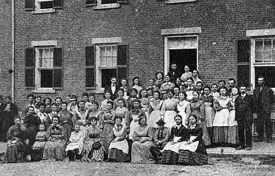 Sepia-toned illustration of young women mill workers standing together outside a brick textile factory in 1830s Lowell, Massachusetts.