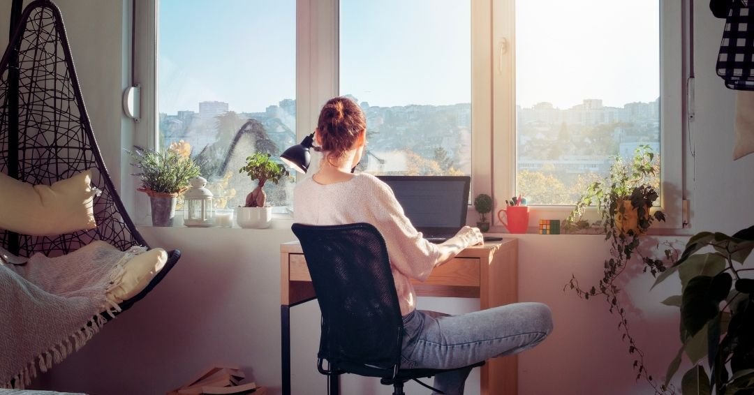 A remote worker at a laptop by a window in a sunlit home office.