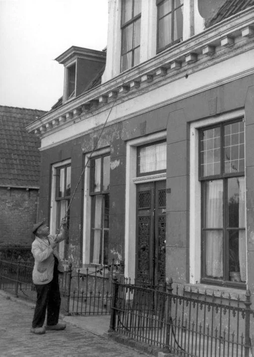A knocker upper man with a long pole tapping on the window of a factory worker to awaken them.