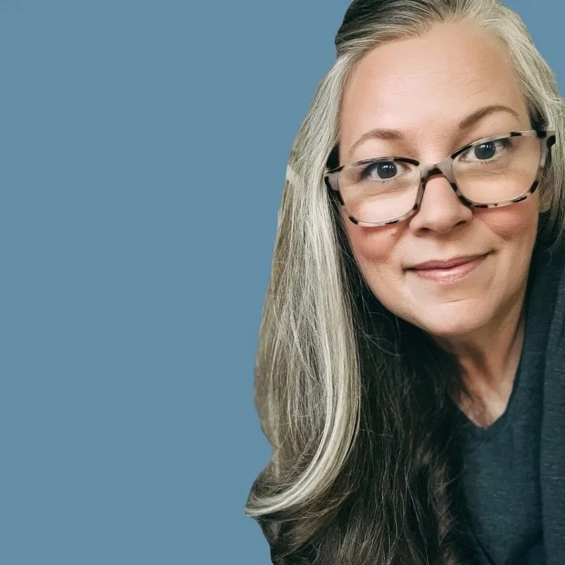 A woman with long, gray and dark hair, wearing glasses and a dark shirt, smiling at the camera against a plain blue background.