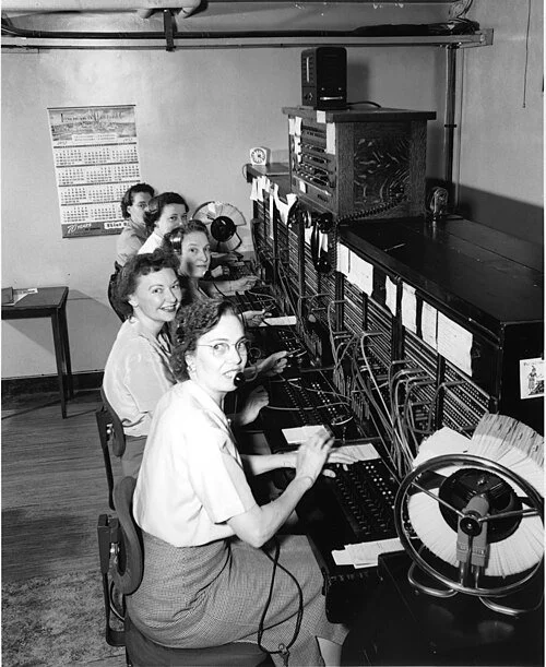 A photograph of a 1950’s telephone switchboard room showing rows of female operators seated at large plug boards, manually connecting calls by inserting cables into jacks to link callers.