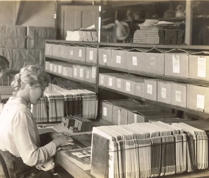 Black and white photograph of an early 1900s factory personnel office with rows of filing cabinets and a clerk at a desk managing worker records.