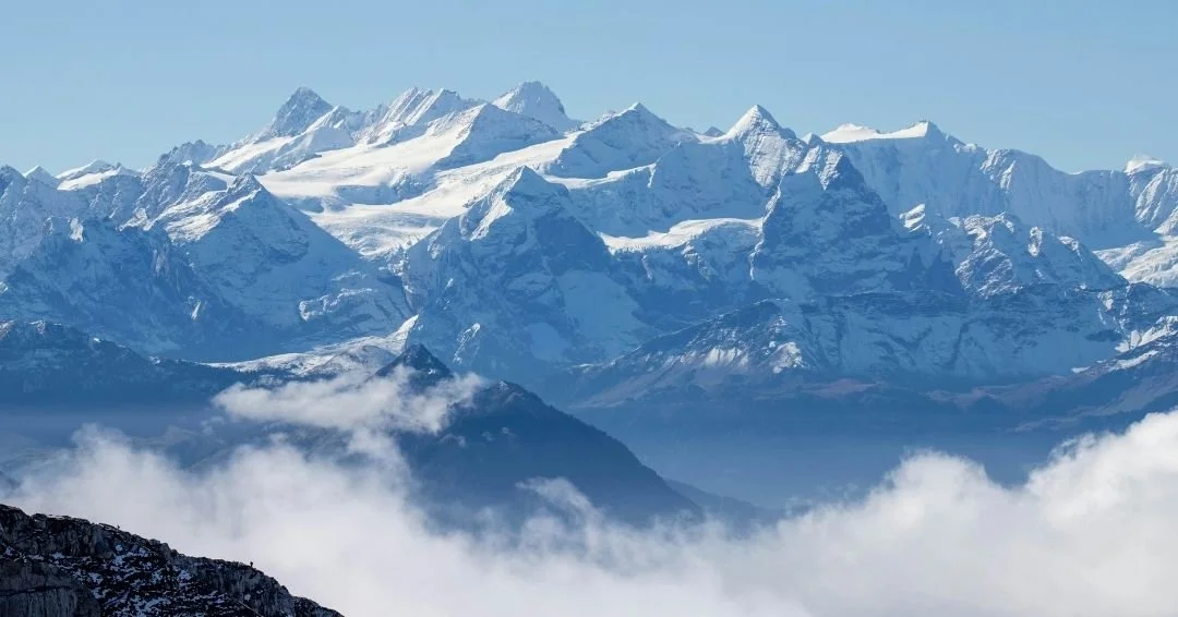 A wide panoramic view of snow-capped mountain peaks rising above a thick layer of white clouds under a clear blue sky. This imagery represents the "summit" of leadership transformation in the History of Work staging area.