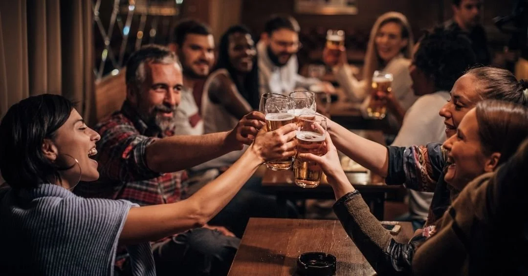 Group of coworkers clinking beer glasses at a bar after work, representing shared peer relationships and informal bonding that can complicate a transition from peer to manager.