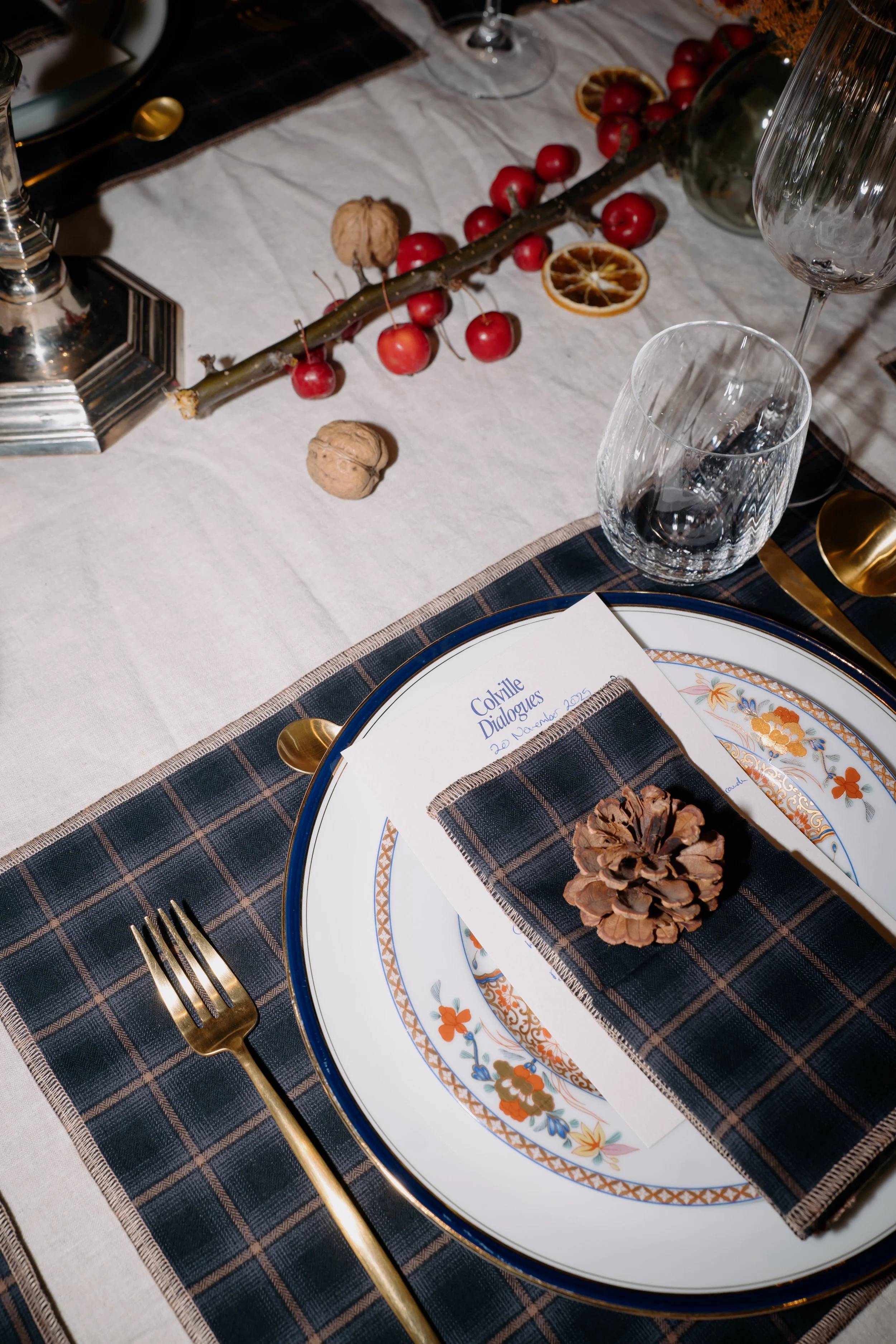 A table setting with a plate and a pine cone on top, a menu or program card, a gold fork, and a gold spoon, set on a dark checkered tablecloth. A transparent water glass and a decorative glass vase are partially visible. The background features a white tablecloth with a branch of red berries, dried orange slices, walnuts, and a candle holder.