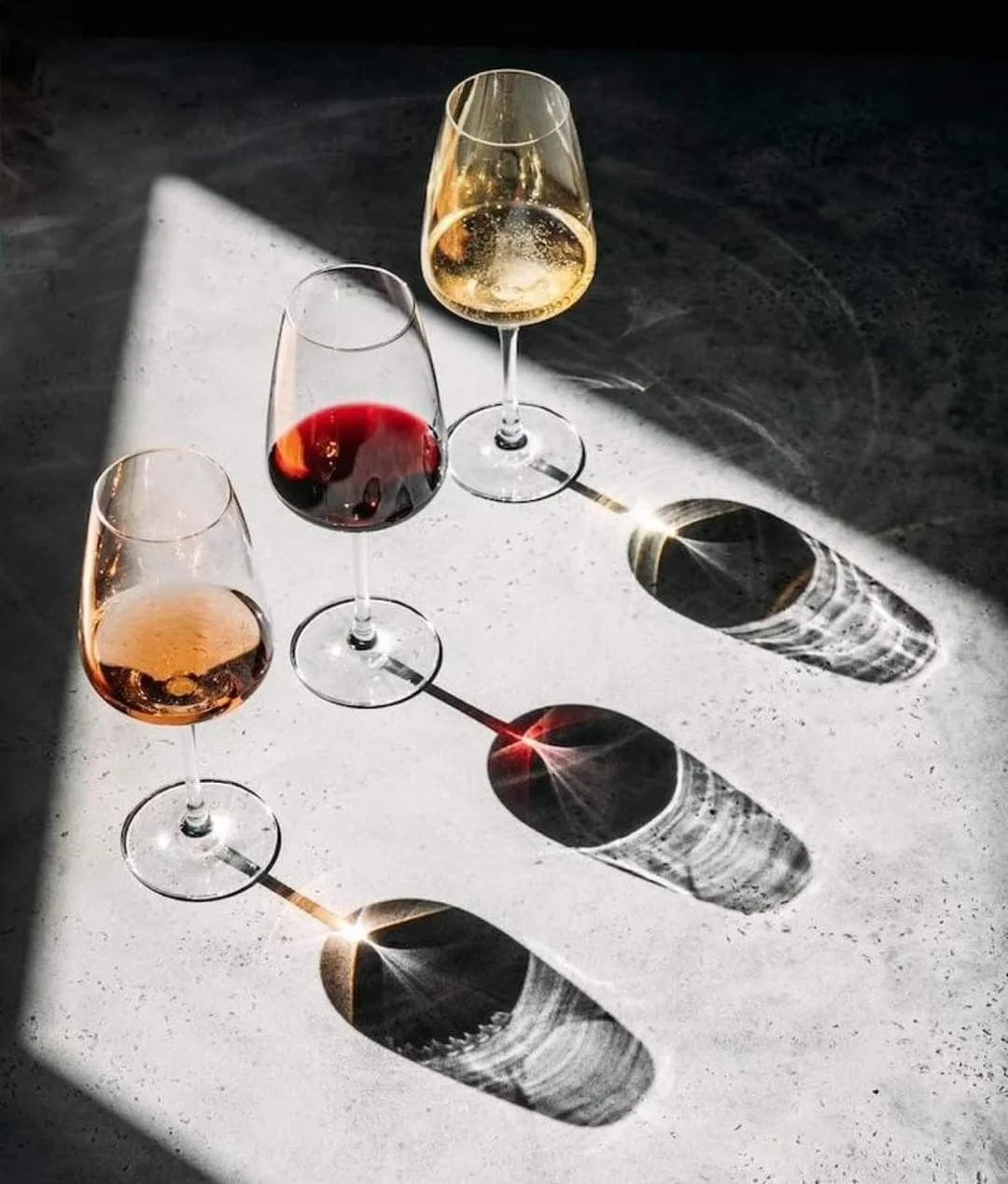 Four glasses of wine on a surface with sunlight casting shadows and reflections.