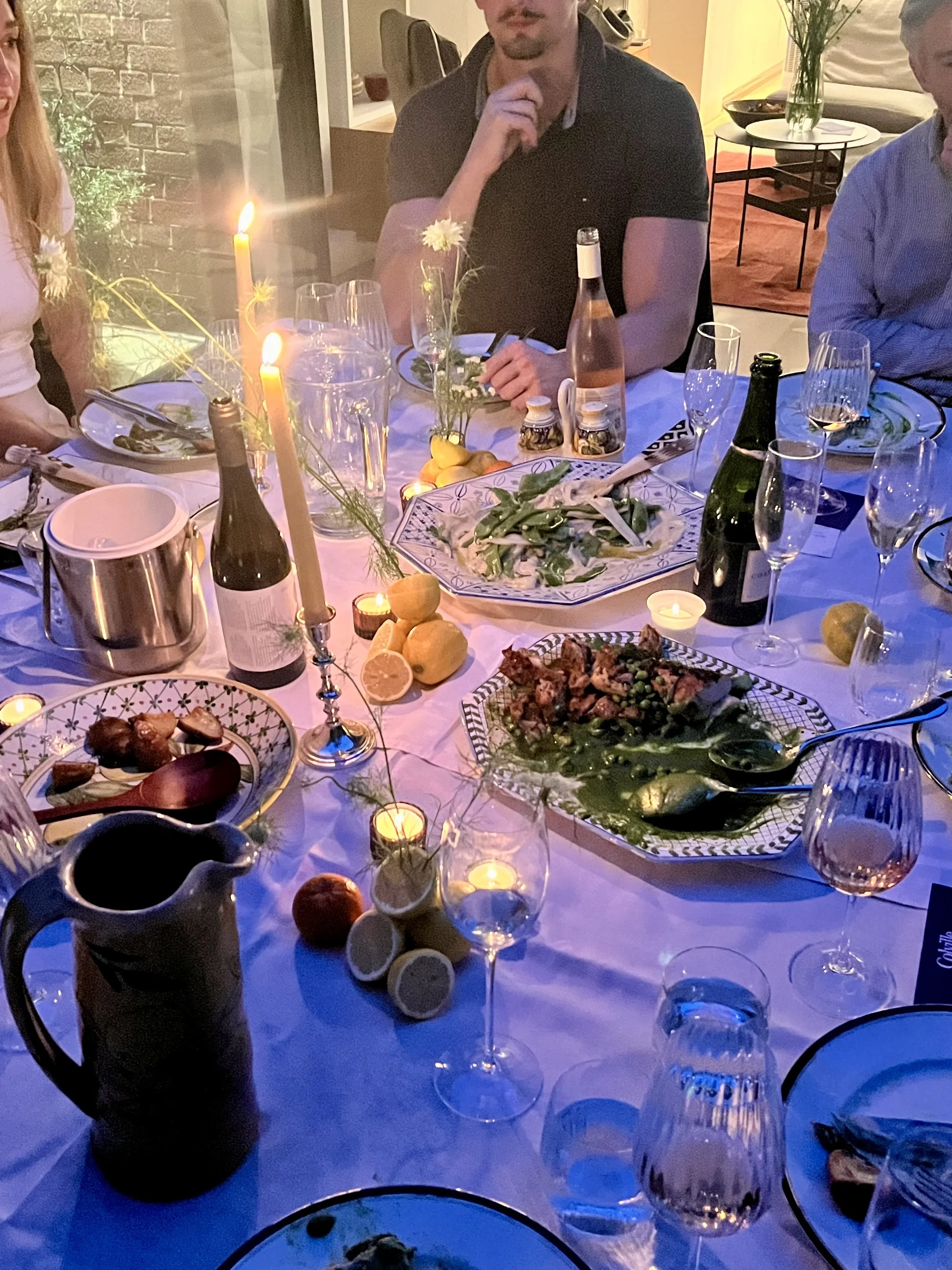 A dinner party scene with a table filled with food, drinks, candles, and floral decorations. Several people are seated around the table, and some are partially visible.