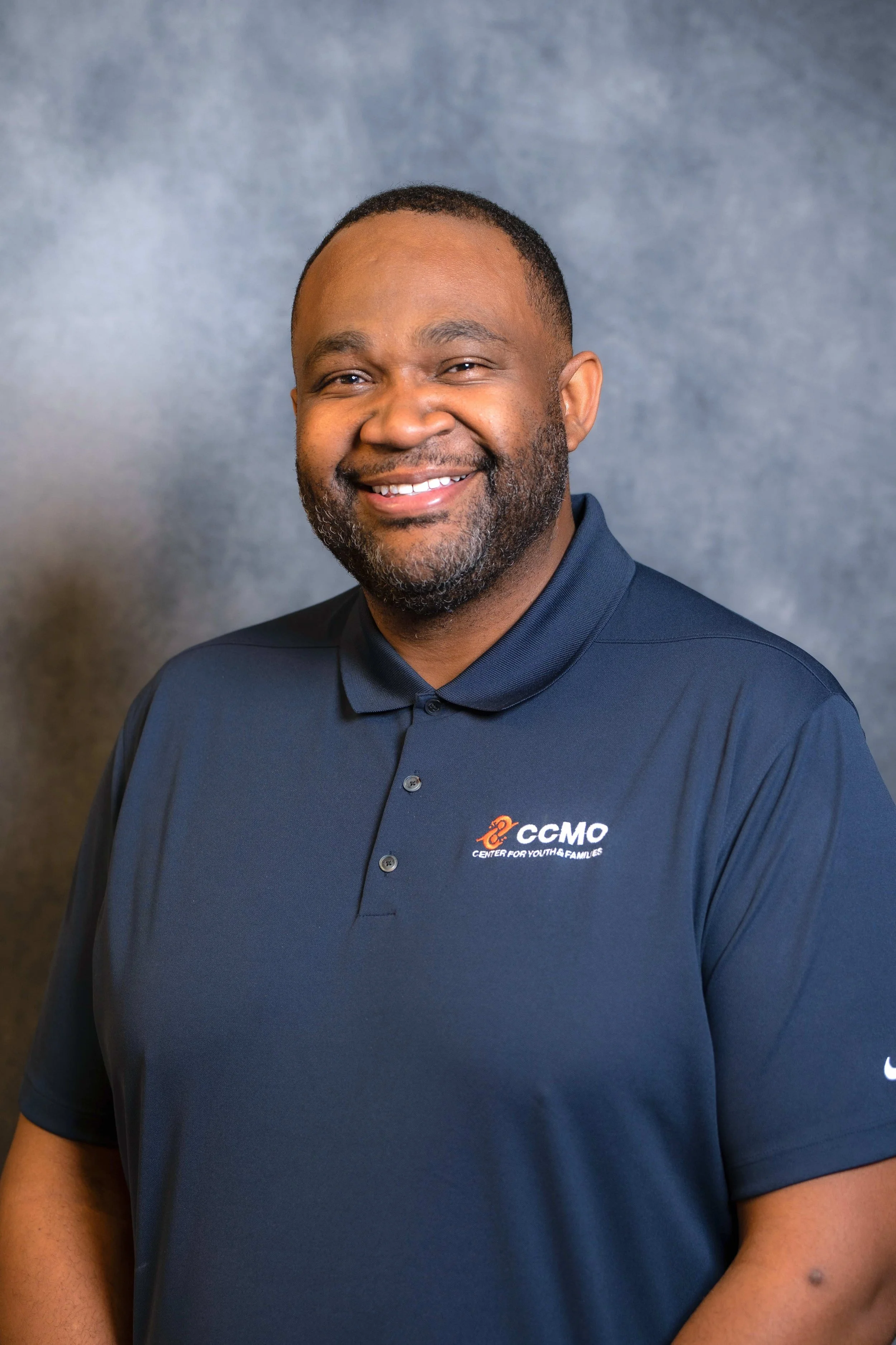 Devin Gough smiling and wearing a navy blue polo shirt, standing against a gray backdrop.