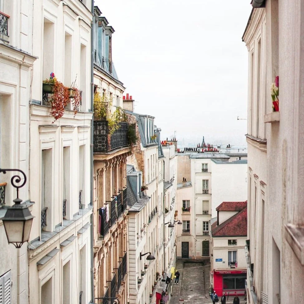 View of a narrow city street flanked by multi-story European-style buildings with balconies, plants, and laundry hanging outside. Some rooftops and the distant horizon are visible in the background.