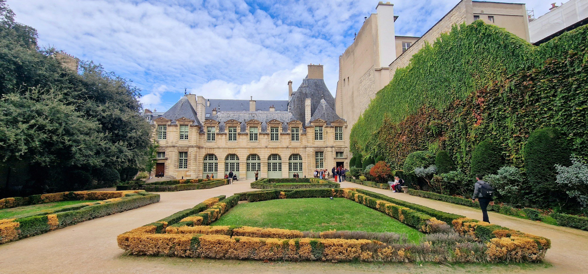 A historic building with large arched windows is surrounded by manicured gardens and a well-maintained walkway, with people strolling and sitting outdoors under a partly cloudy sky on a French Voyage planned tour.