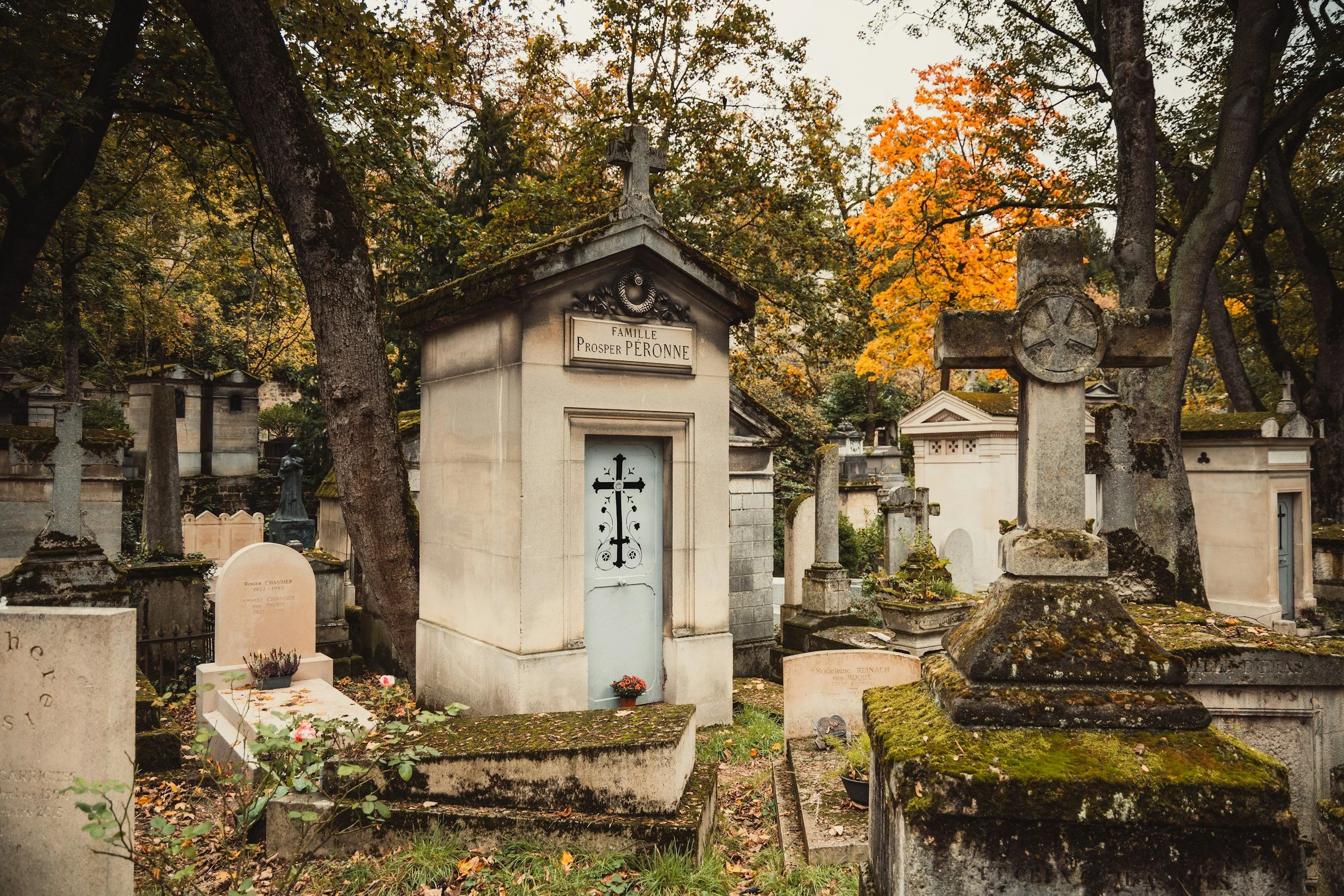 Old cemetery with various weathered tombstones and crosses, surrounded by trees with autumn foliage, for a French Voyage planned private tour of Pere Lachaise Cemetery.