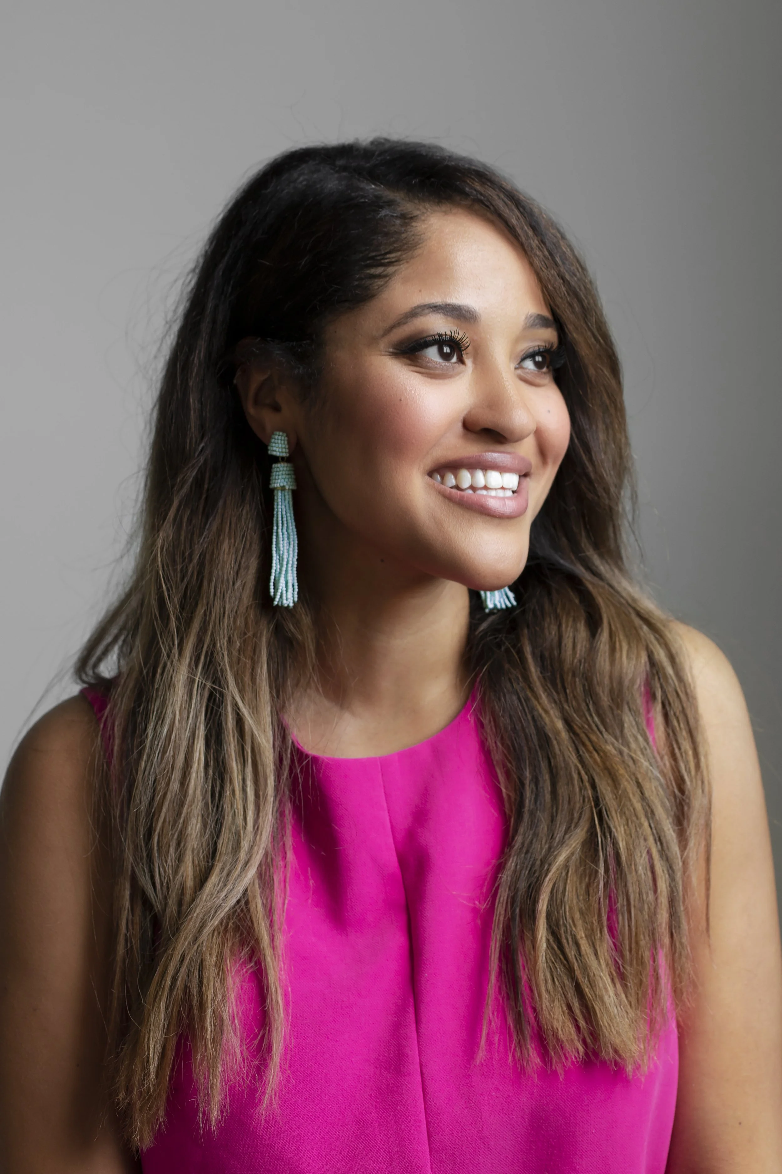 French Voyage CEO & Founder, Monique Malone - a woman with long brown hair, wearing a bright pink sleeveless top and turquoise beaded earrings, smiling and looking slightly to her right against a gray background.