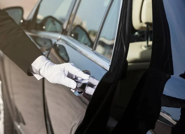 A private driver or chauffeur wearing a white glove opening the door of a black luxury car for a French Voyage client.