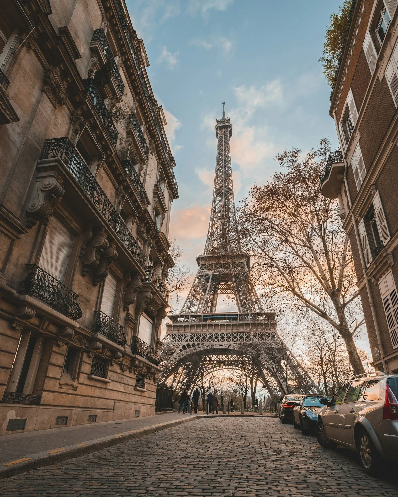 View of the Eiffel Tower in Paris, France, with surrounding historic buildings, cobblestone street, some parked cars, and leafless trees during sunset or early evening.