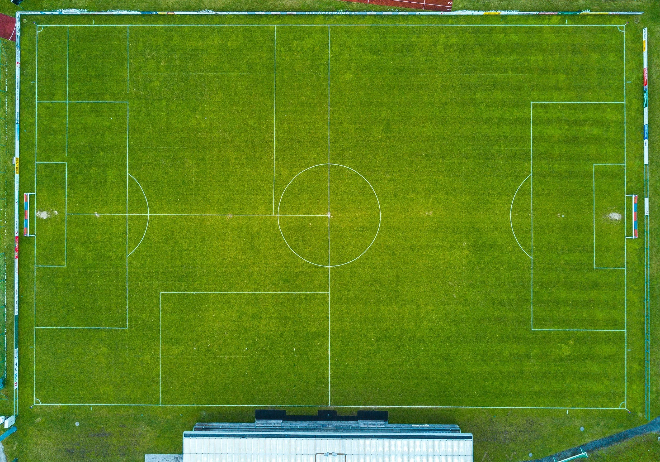 Aerial view of a grass pitch with green grass, white lines marking the boundaries, center circle, penalty areas, and goals at each end.