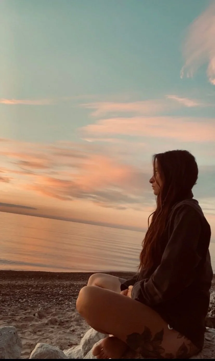 A woman sitting cross-legged on a rocky beach during sunset, facing the water with colorful clouds in the sky.