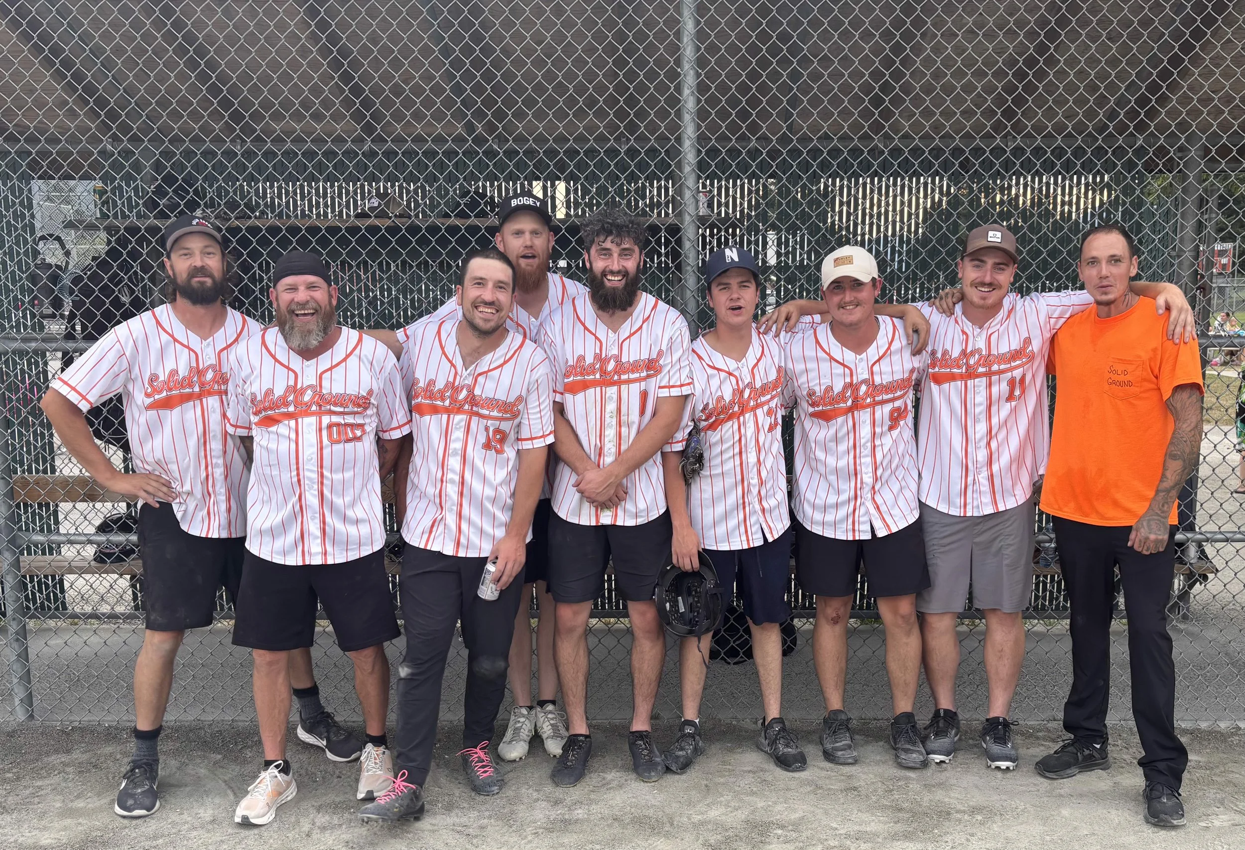 Group of nine men in baseball uniforms and one man in an orange shirt standing in front of a chain link fence at a baseball field, smiling and posing for a photo.