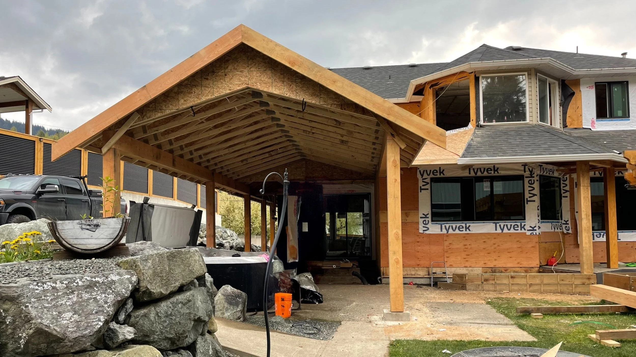 Front view of a house under construction with a wooden porch and deck, windows, and outdoor area, with construction materials and vehicles nearby.