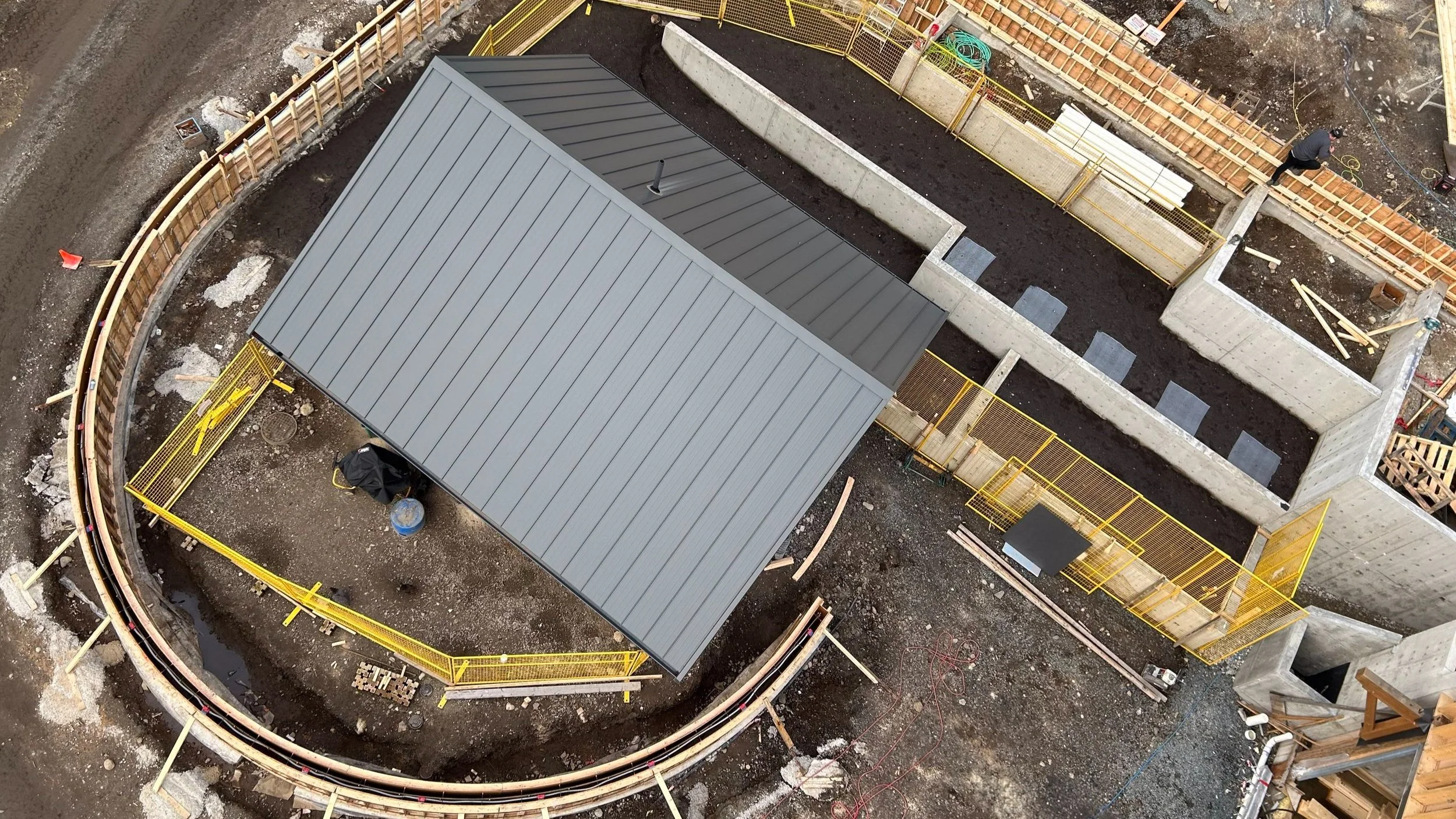 Construction site viewed from above showing a newly installed gray metal roof over a small building, surrounded by dirt, architectural concrete, and yellow safety fencing, with workers in the background.