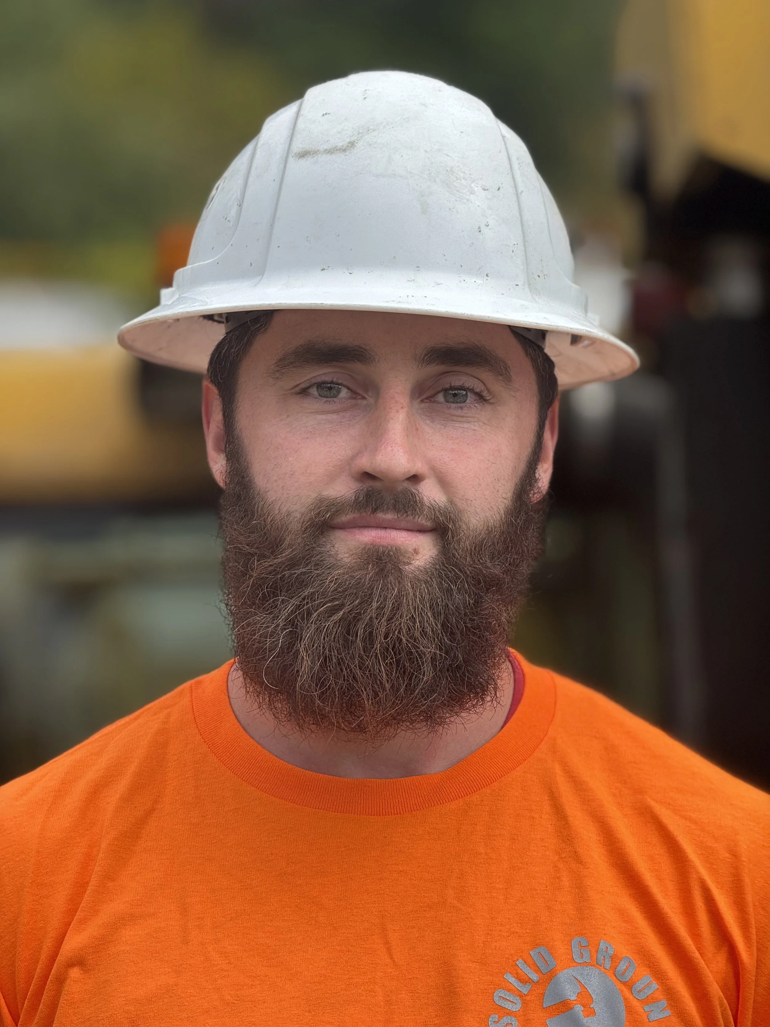 A man with a full beard and light-colored eyes wearing a white construction helmet and an orange t-shirt with a logo that says 'Solid Ground'.