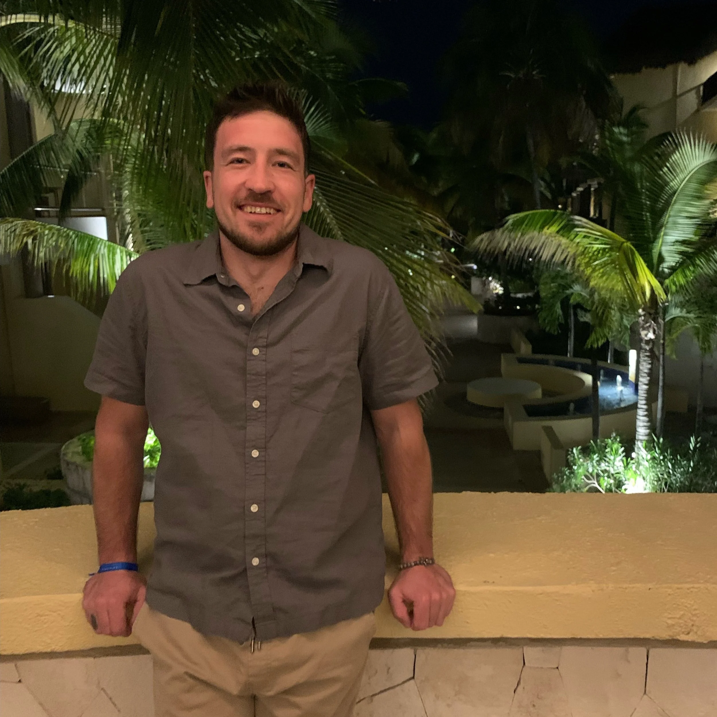 A man with short dark hair and a beard smiling, leaning on a yellowish bar or wall outdoors at night, with large green palm trees and decorative lighting in the background.