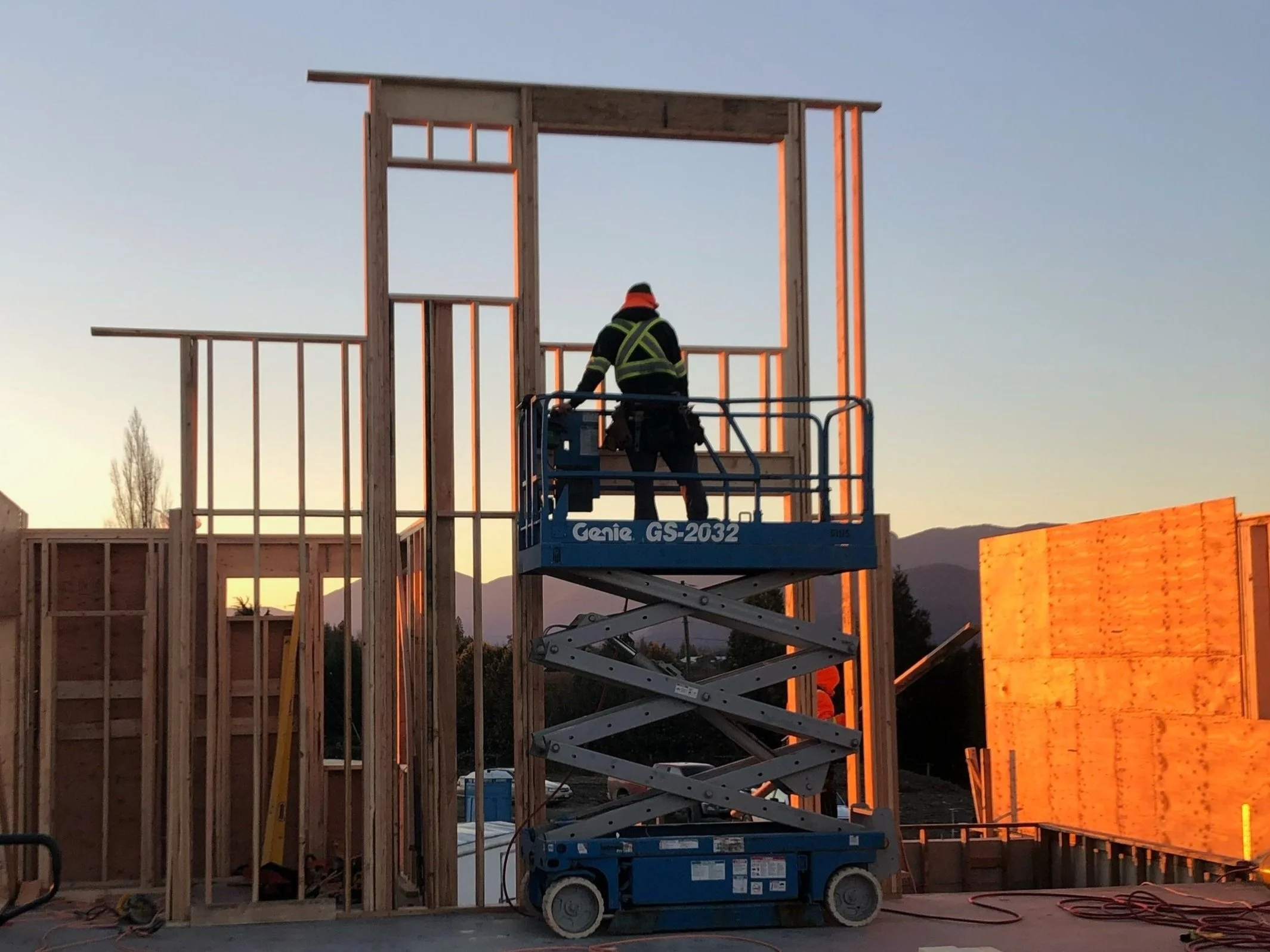 Construction worker on a blue scissor lift installing wooden framing for a building at sunset with mountains in the background.