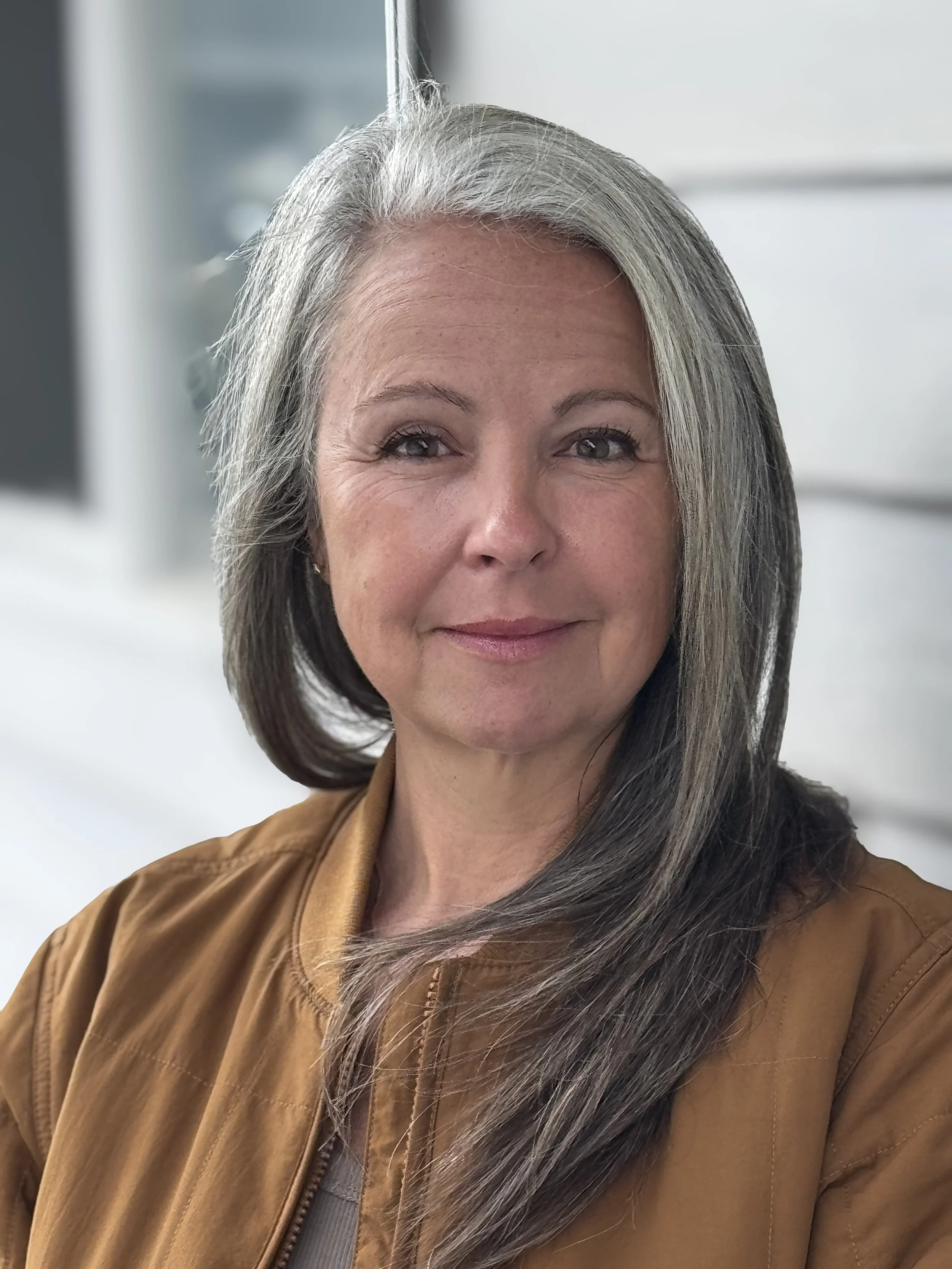 Portrait of a middle-aged woman with long gray hair, smiling, wearing a brown jacket, indoors near a window.