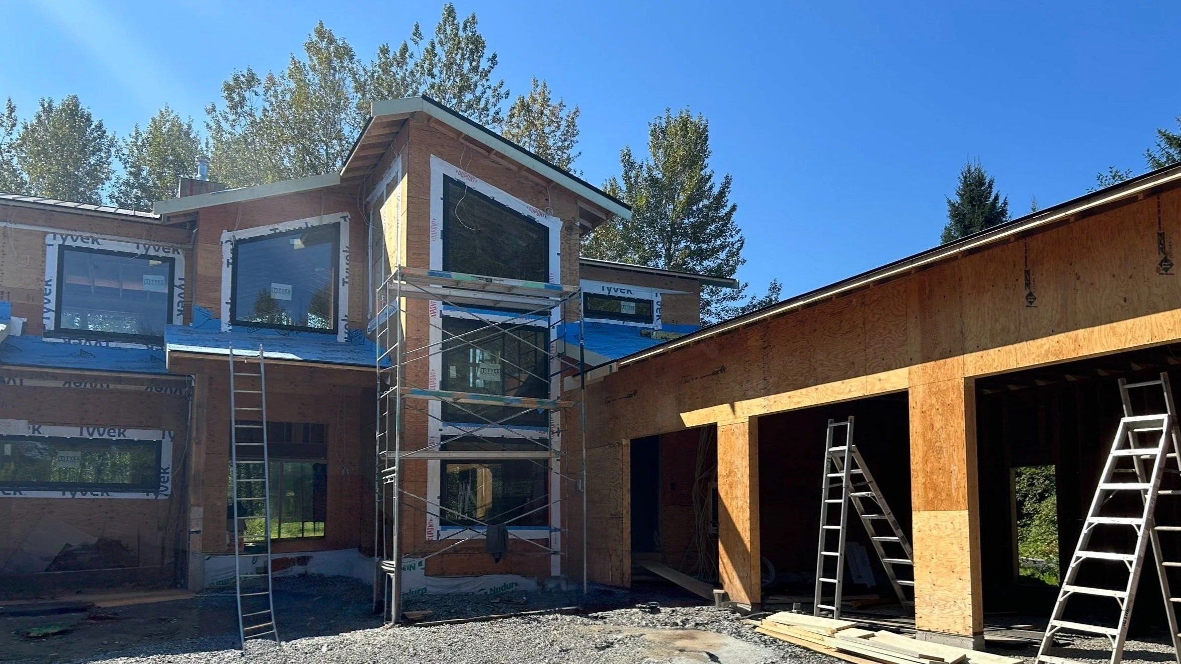 Construction site of a house with wood framing and scaffolding, blue sky, and trees in the background.