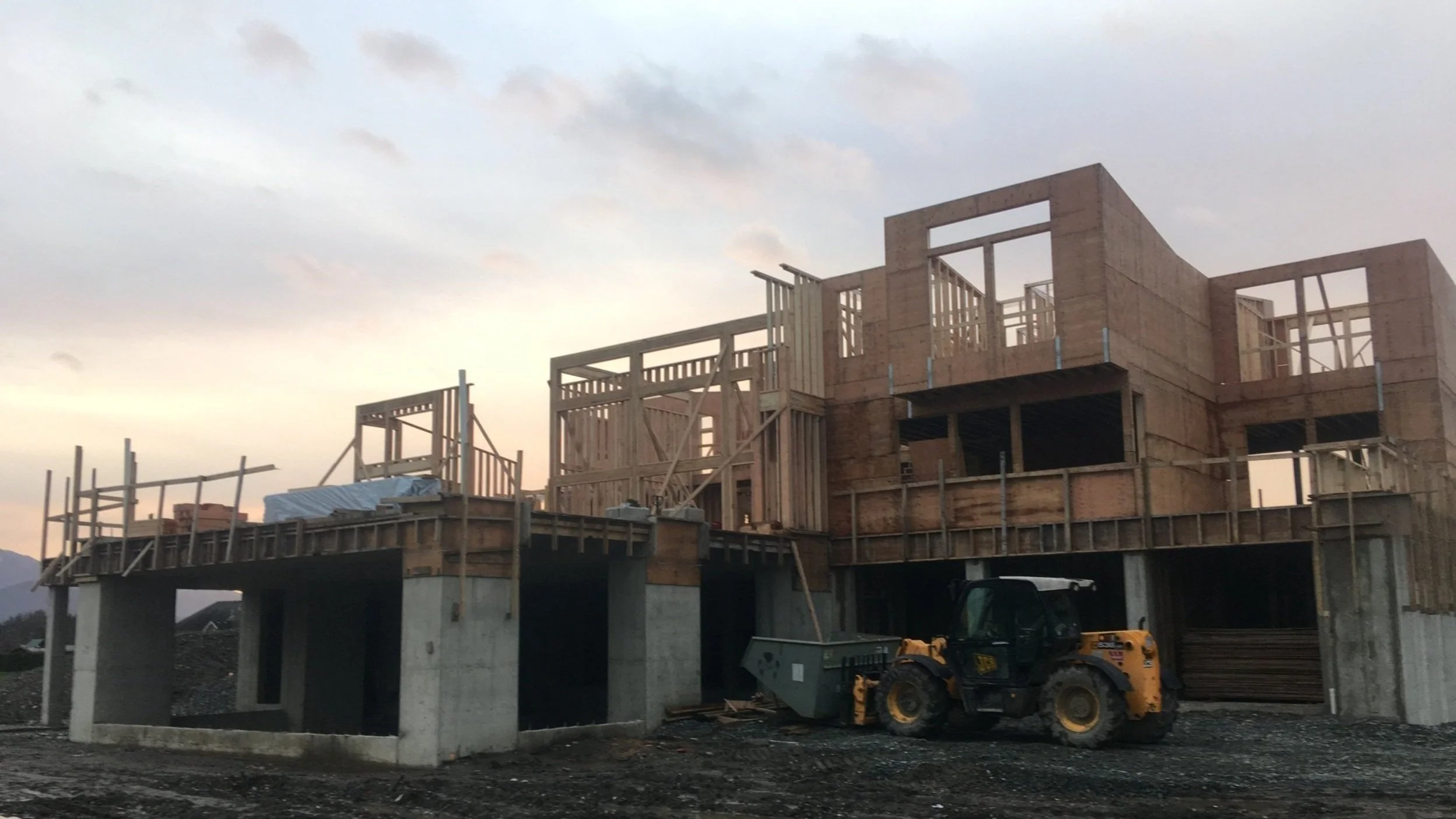 Under construction wooden house framework on concrete foundation with construction equipment nearby at sunset