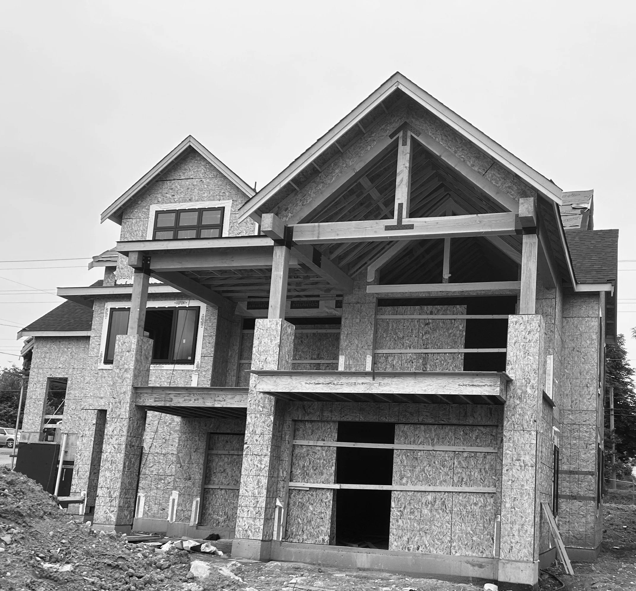 Black and white photo of a house under construction, showing an incomplete structure with exposed wood and plywood sheathing