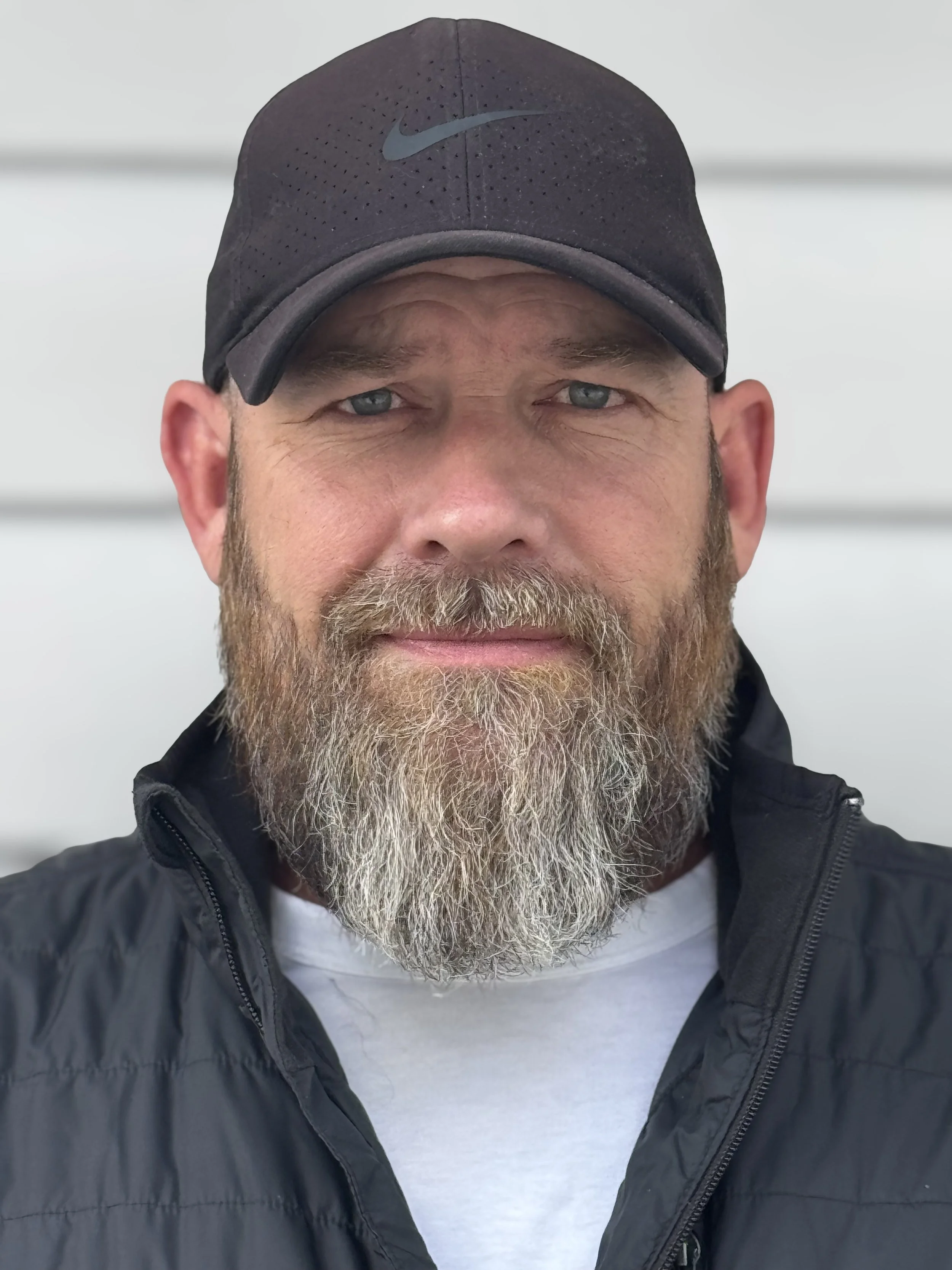 A man with a full gray beard wearing a black Nike cap, a black jacket, and a white t-shirt standing in front of a white wall.