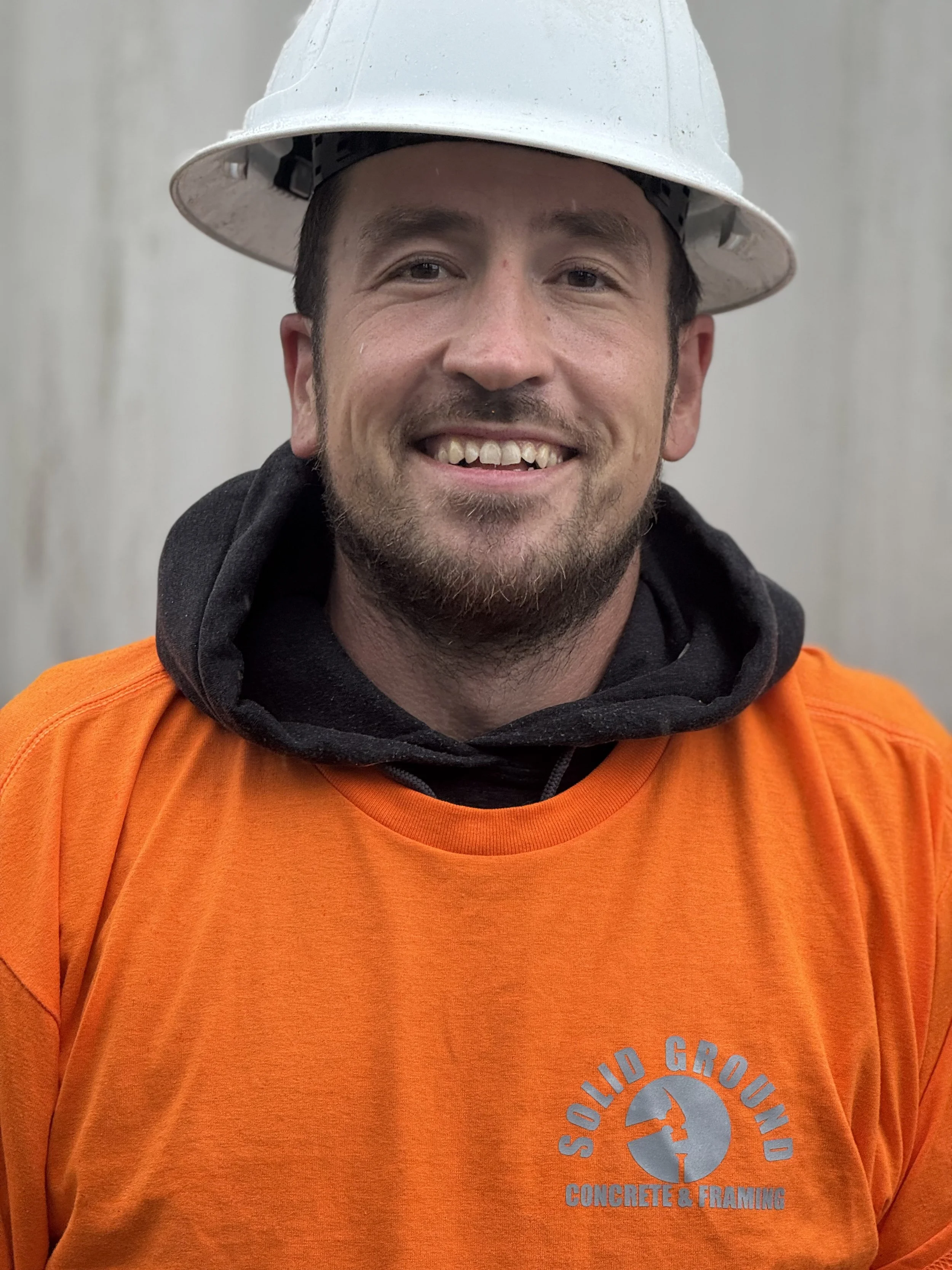 A smiling man wearing a white safety helmet, black hoodie, and bright orange shirt with 'Solid Ground Concrete & Framing' logo.