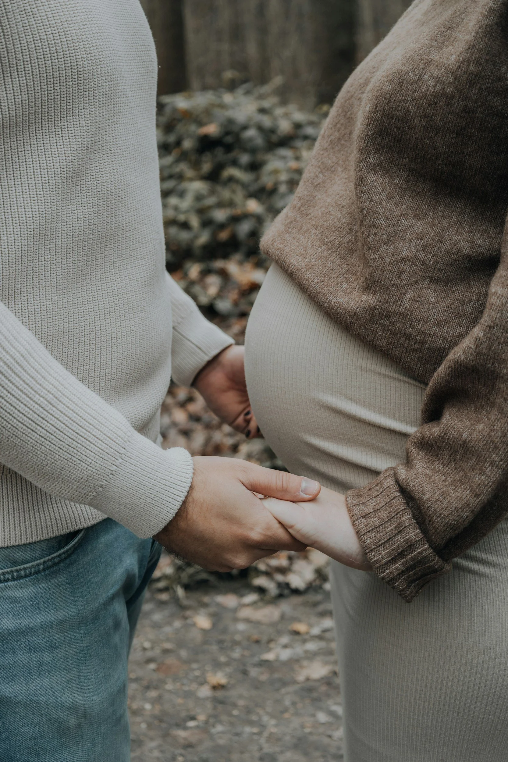A couple holding hands, with the woman pregnant, outdoors with trees and fallen leaves in the background.