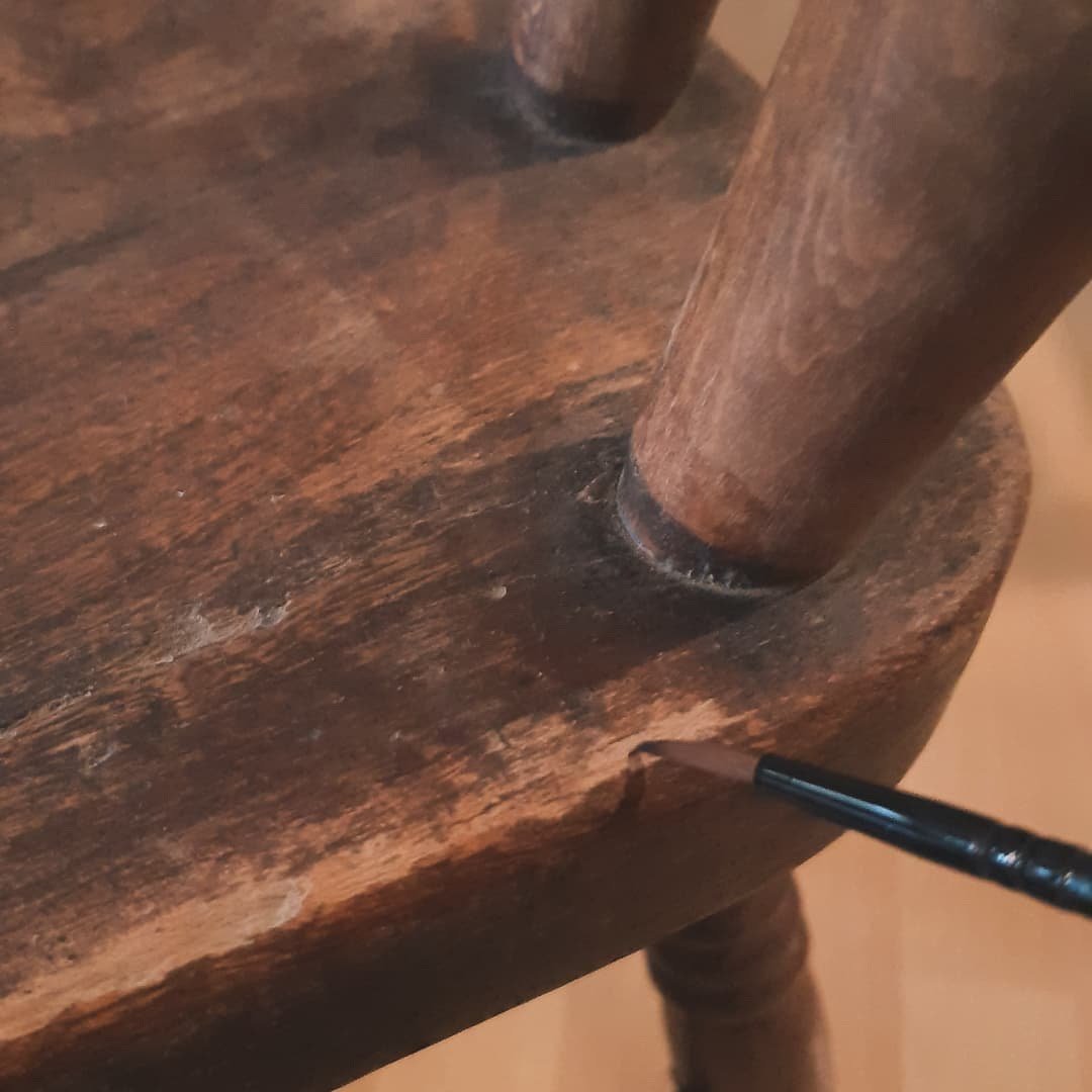 Detailaufnahme einer Stuhl-Holzoberfläche bei der Möbelaufarbeitung, mit Pinsel wird eine abgenutzte Stelle am Möbelstück ausgebessert.