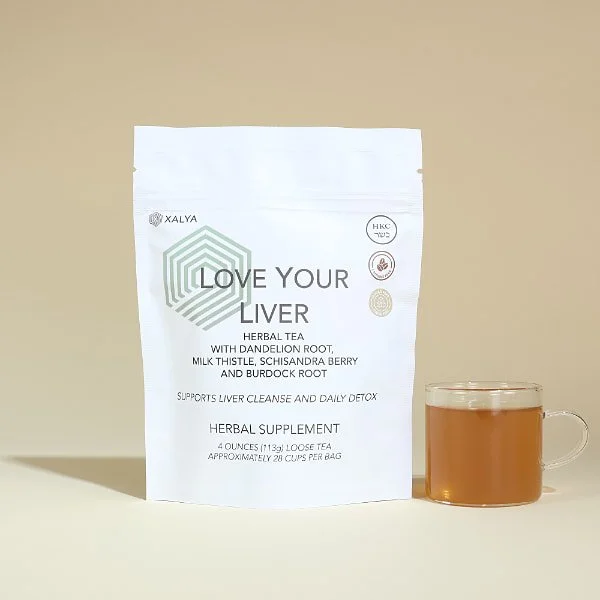 A white stand up pouch of XALYA Love Your Liver herbal detox tea next to a glass mug of amber-colored brewed tea.