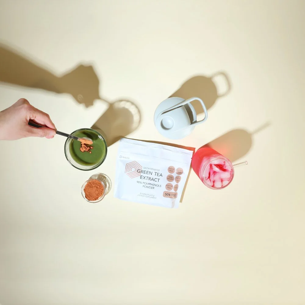 Overhead view of XALYA Green Tea Extract Powder pouch with a spoon adding powder to a green smoothie drink, alongside a glass of pink iced beverage, a shaker bottle, and a small bowl of powder on a beige background.