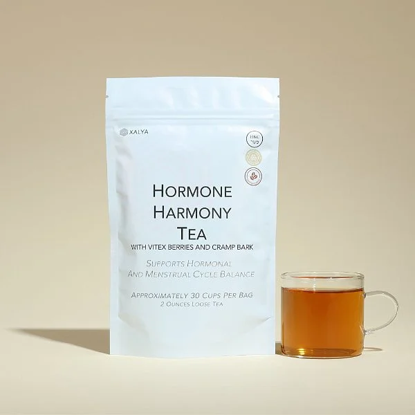 A white stand-up pouch labeled "XALYA Hormone Harmony Tea" with berries and bark, next to a glass mug filled with tea.
